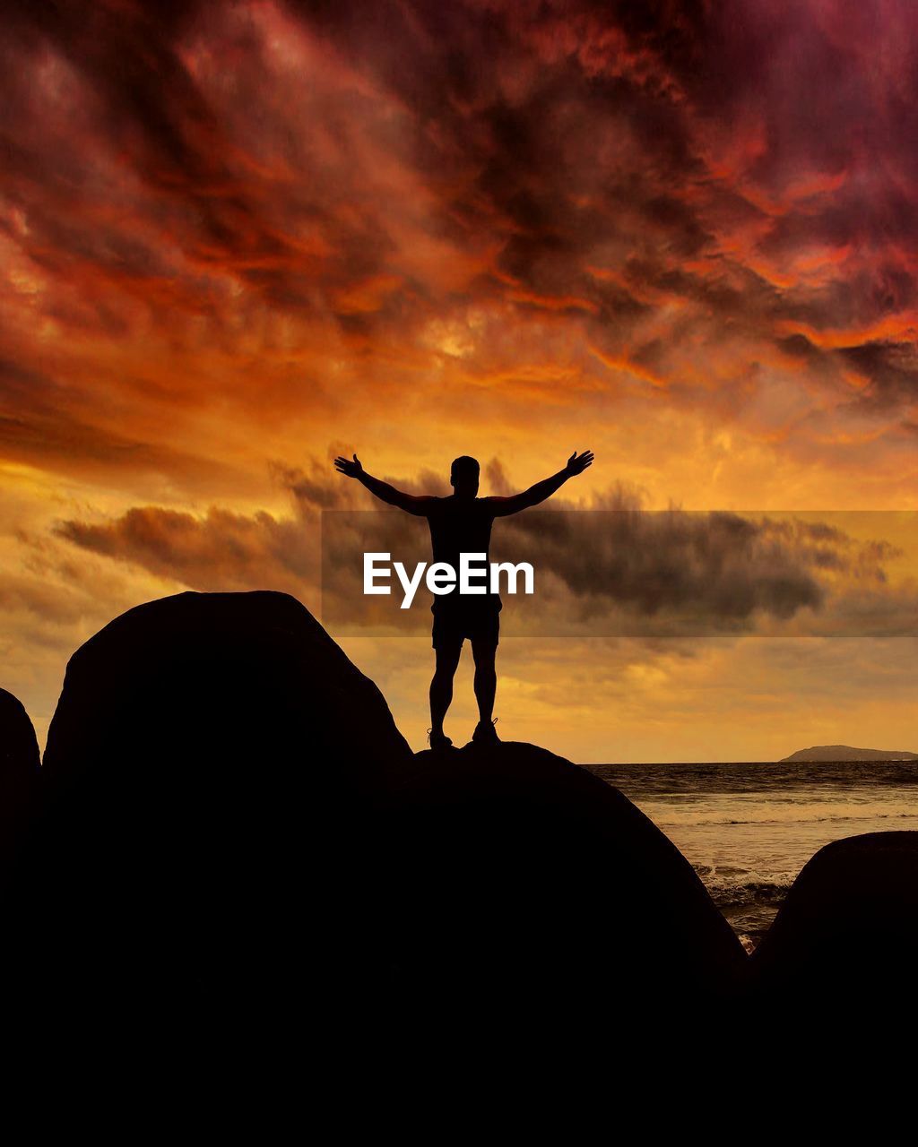 Rear view of silhouette man with arms outstretched standing on rock by sea against sky during sunset