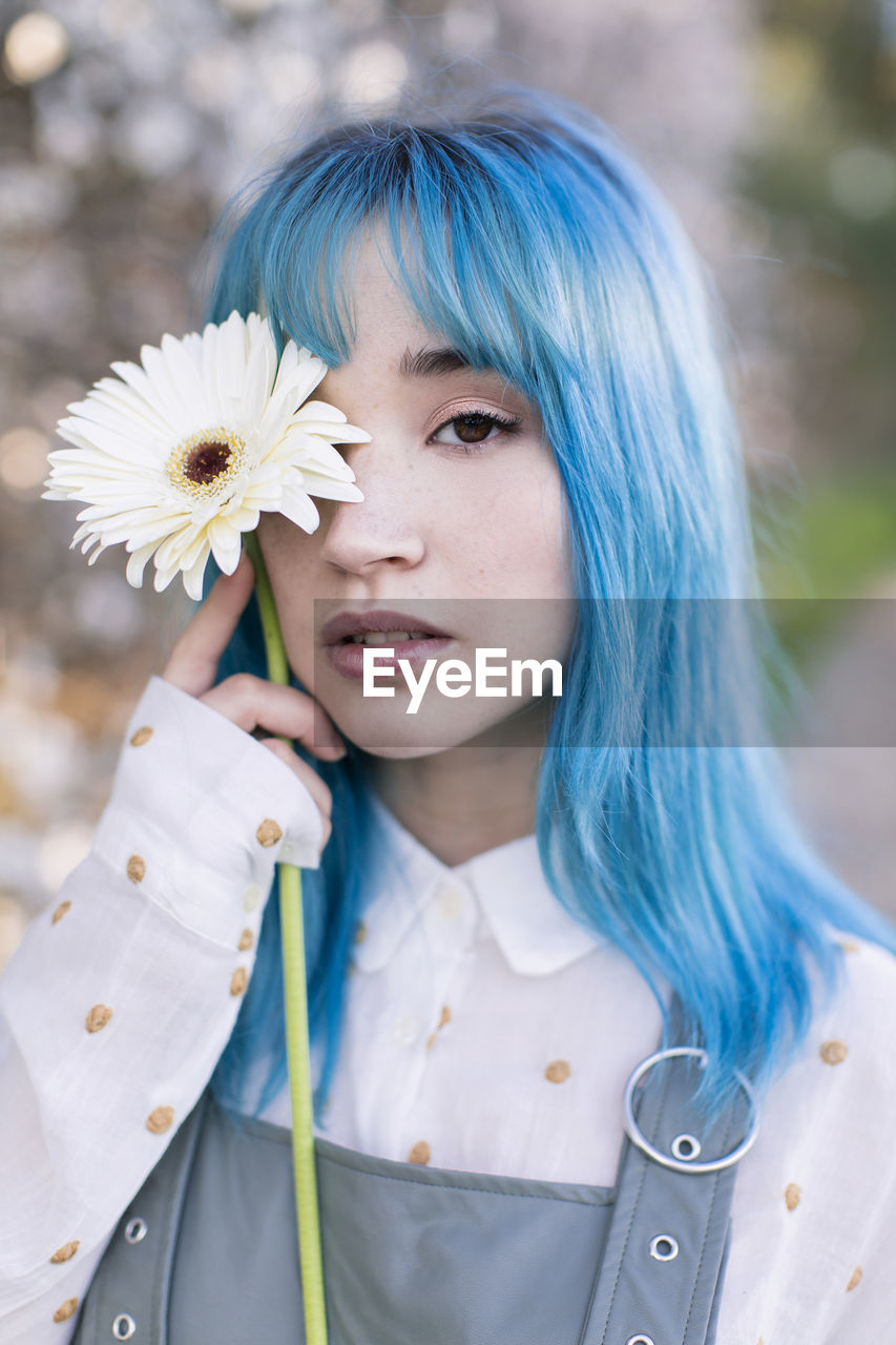 Modern trendy female with blue hair holding a fresh flower covering eye and looking at camera while standing in blooming spring garden