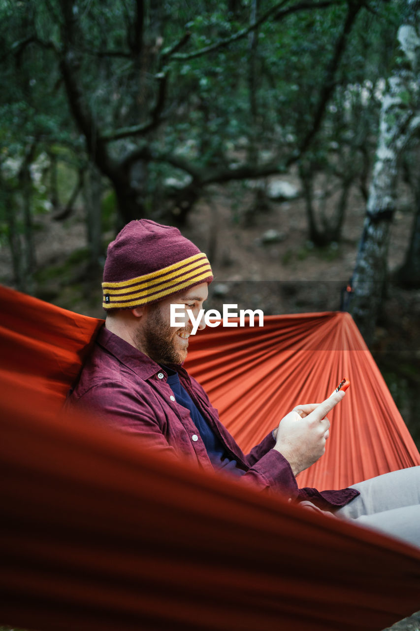 Young male uses his smartphone resting on a hammock in the woods