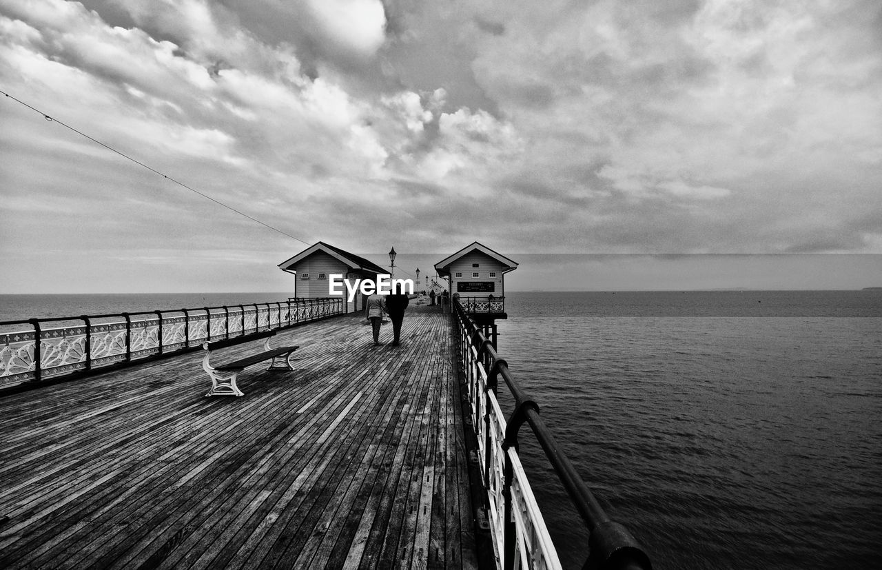 Man and woman walking on bridge over sea against cloudy sky