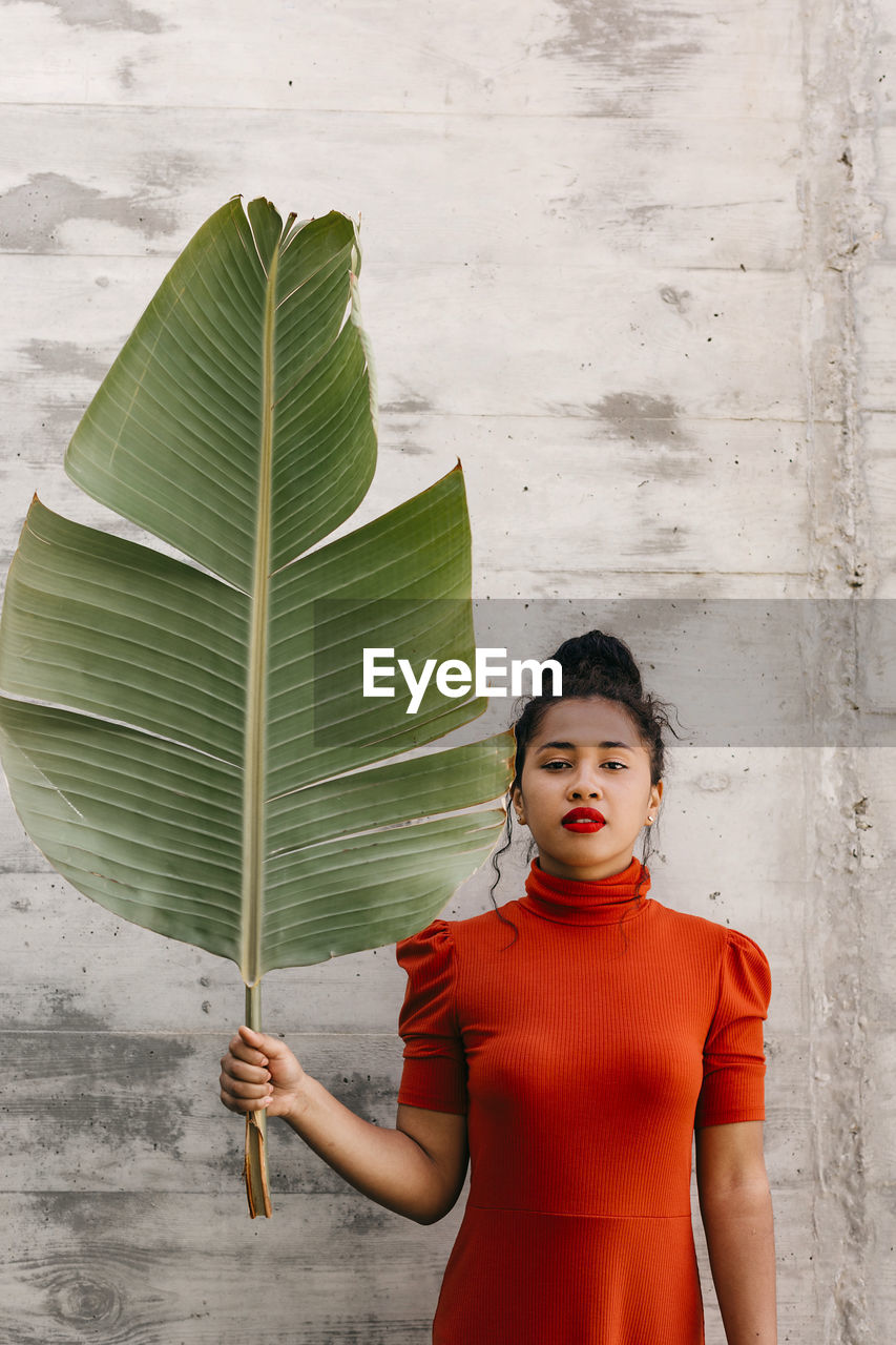 Young woman in turtleneck red dress holding green banana leaf in front of wall