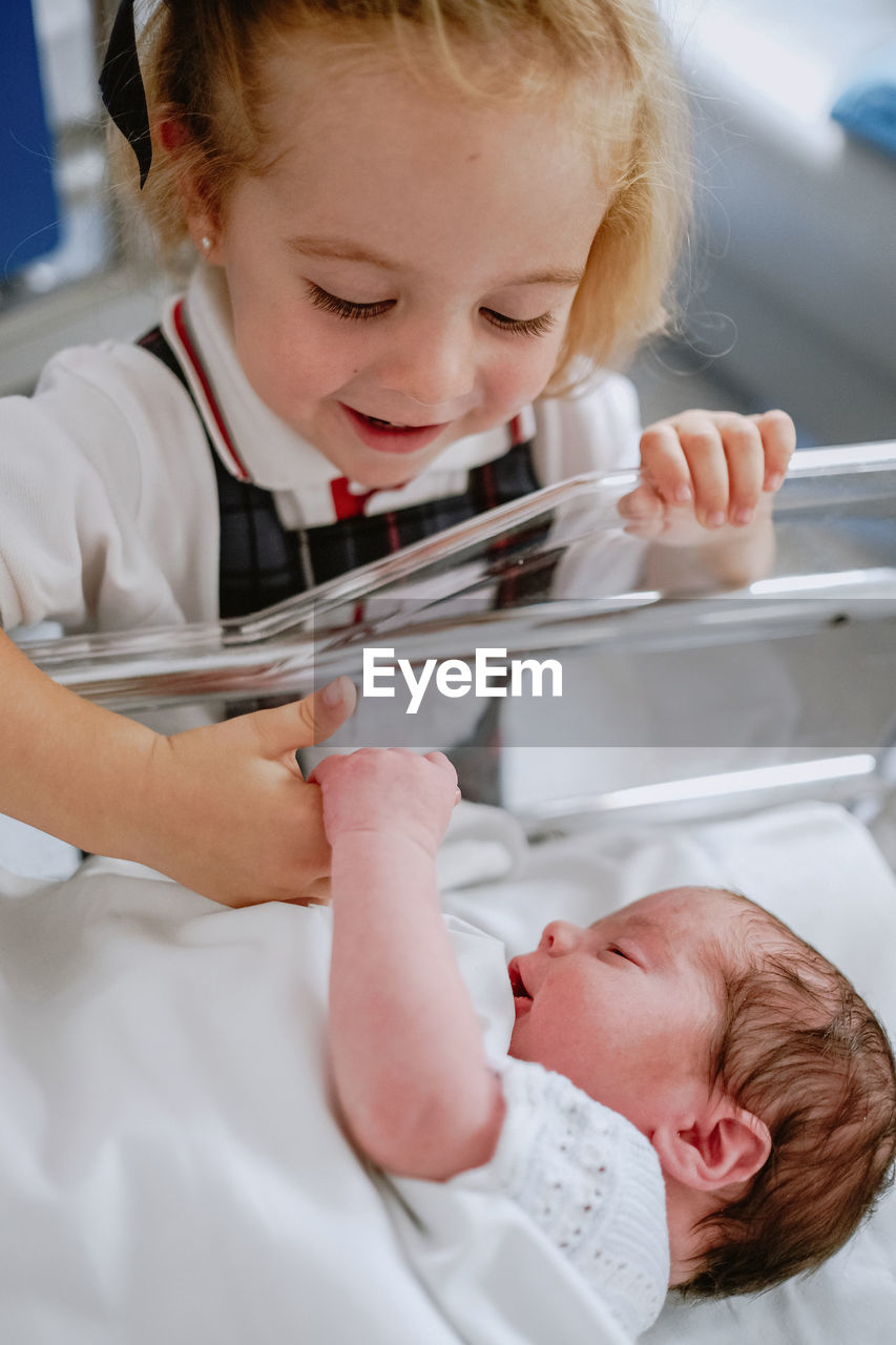 From above of cheerful curious little girl standing near newborn baby sibling sleeping in hospital cradle in ward