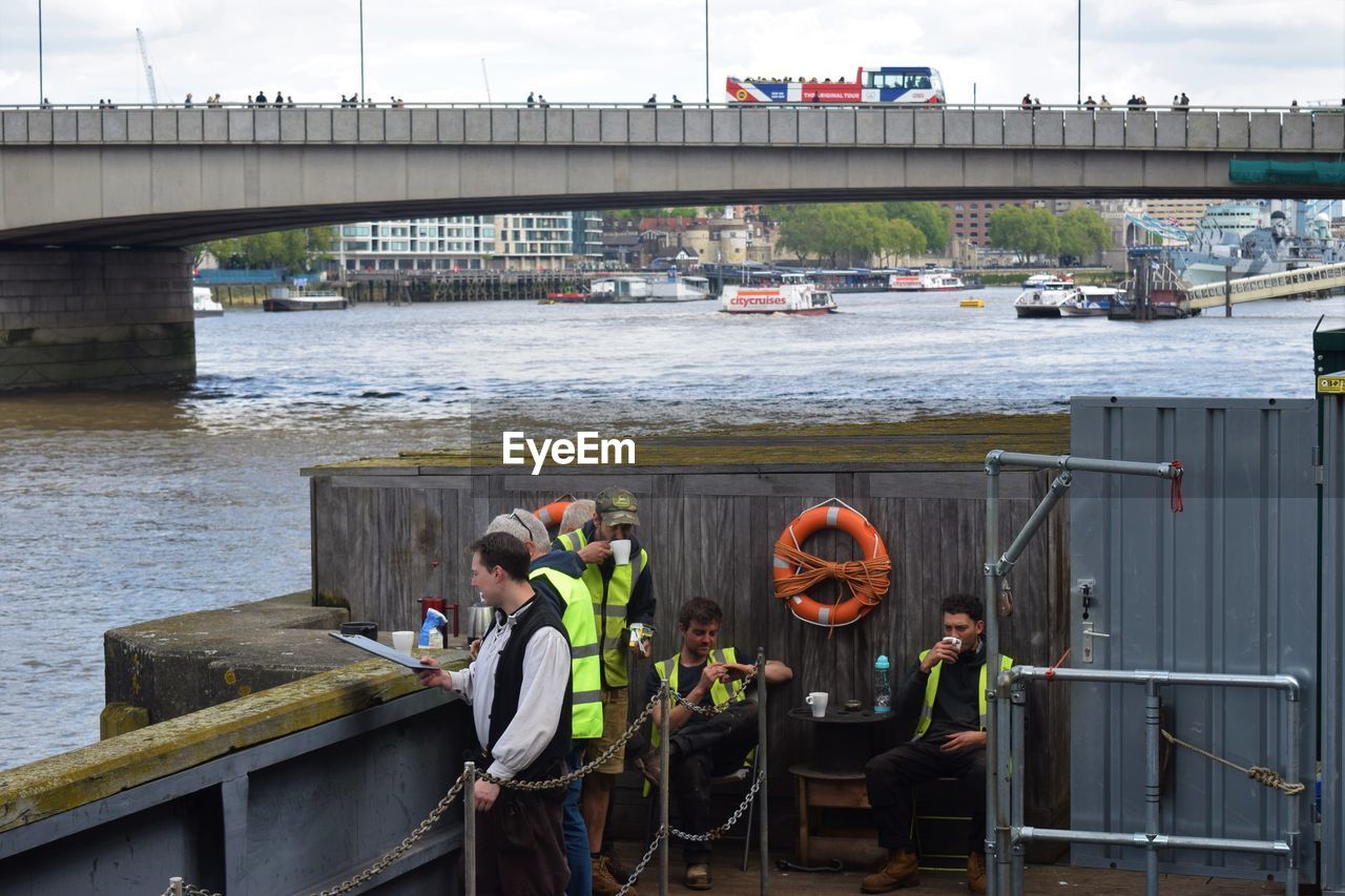 PEOPLE STANDING BY BRIDGE OVER RIVER AGAINST SKY