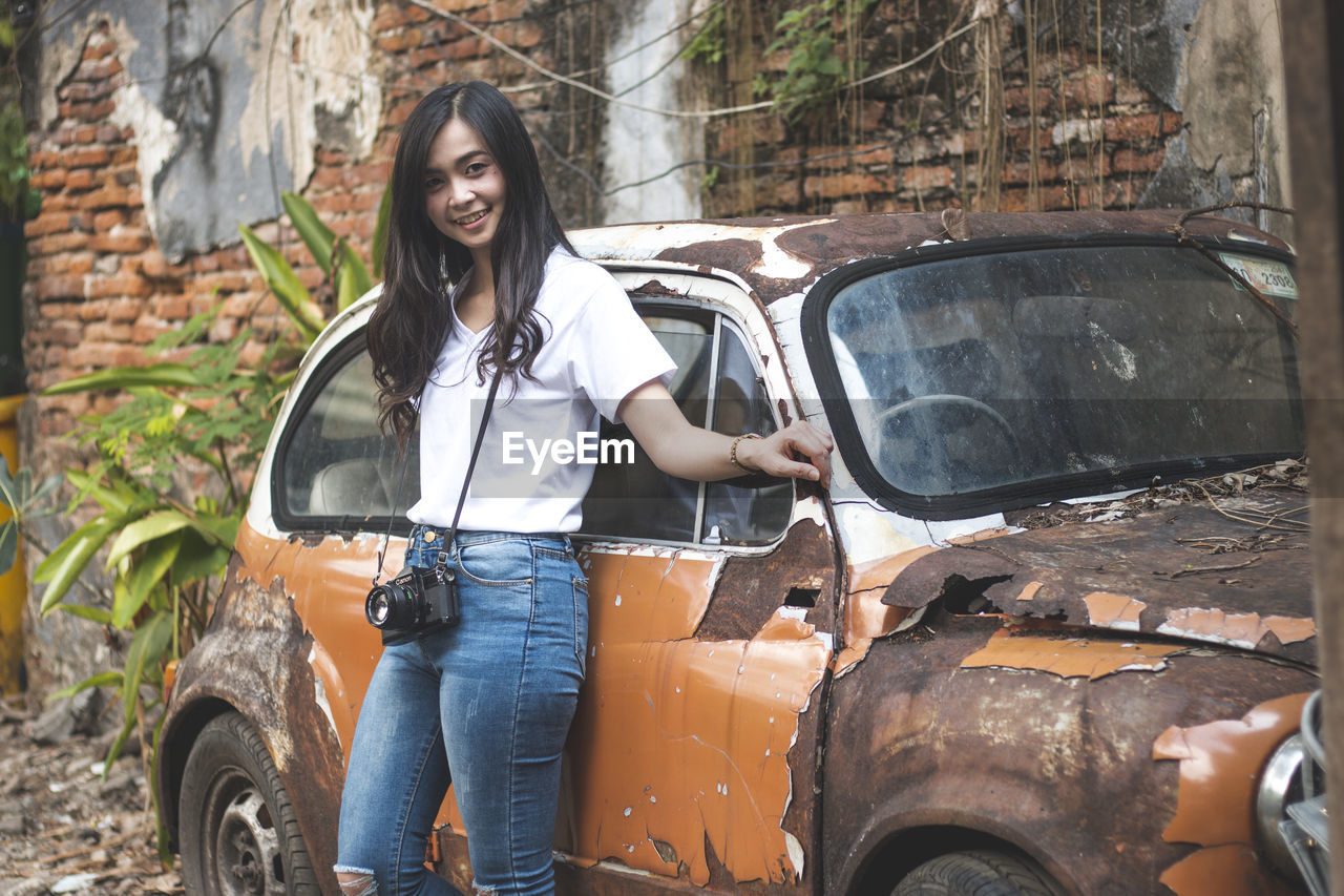 Portrait of smiling young woman standing by abandoned car