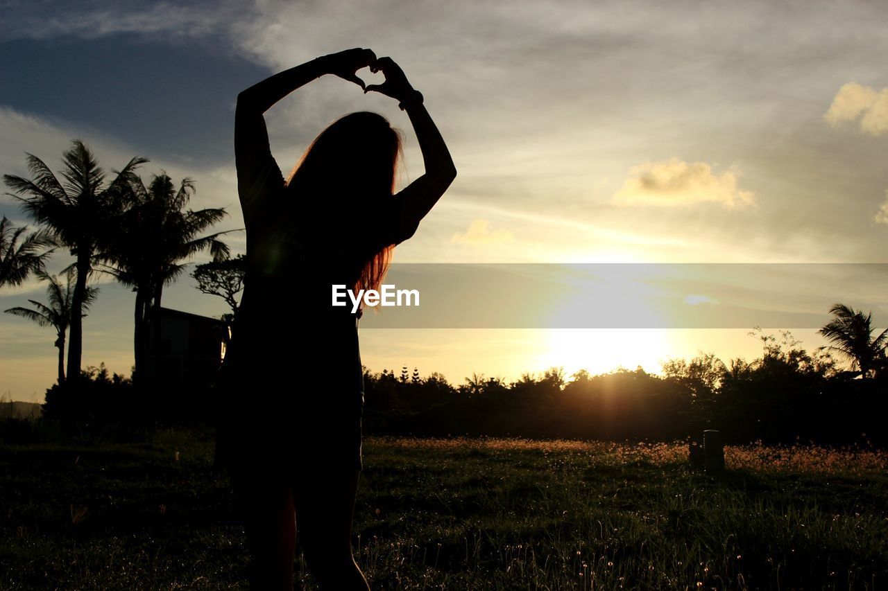 Silhouette woman making heart shape with hands on grassy field against sky during sunset
