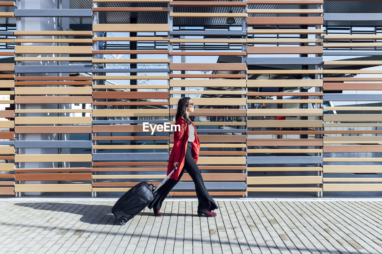 Businesswoman walking on footpath with suitcase by wooden wall during sunny day