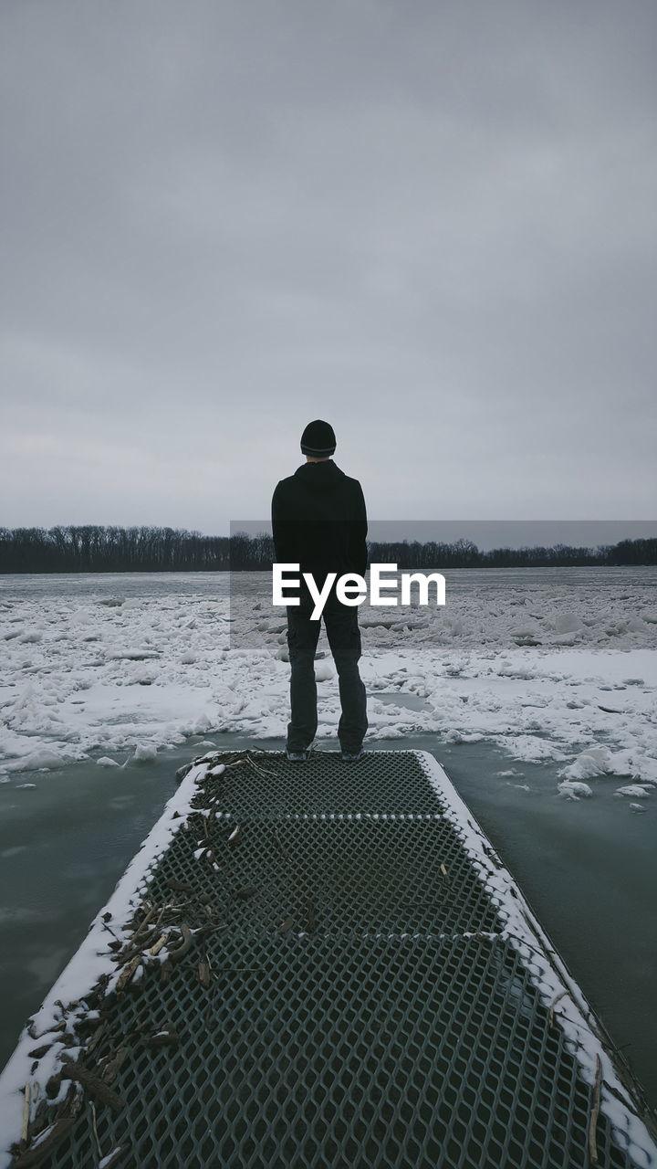 Rear view of man standing by frozen lake against cloudy sky