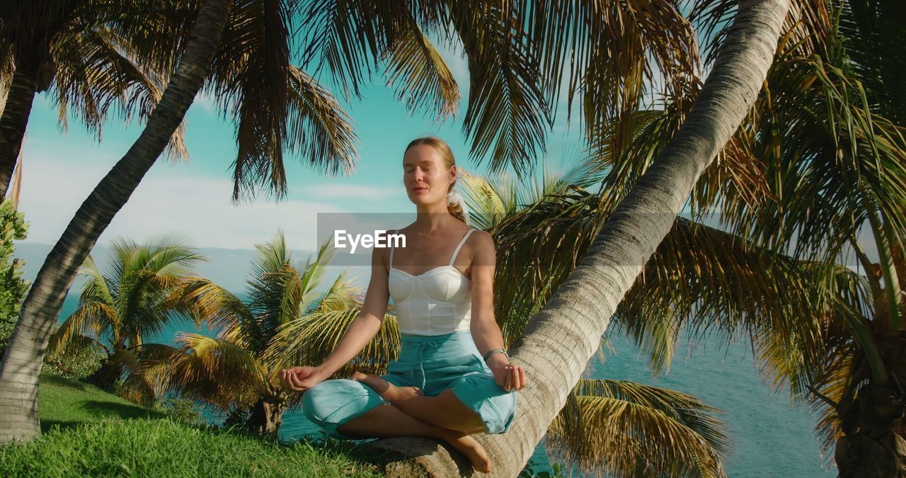 Girl meditates on grassy lawn in a lotus position and breathes deeply. tropical palm trees on ocean.