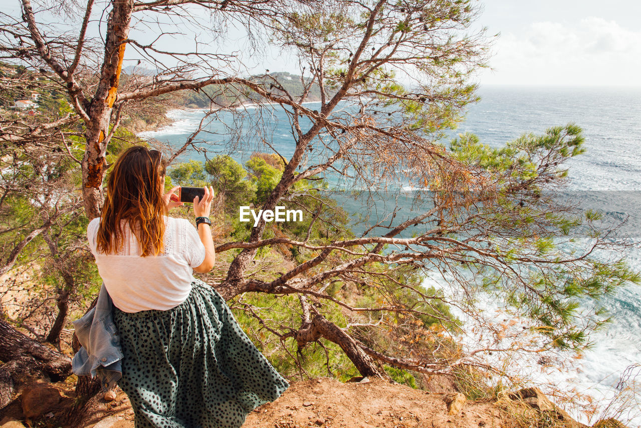 Woman photographing sea with smart phone while sitting on tree