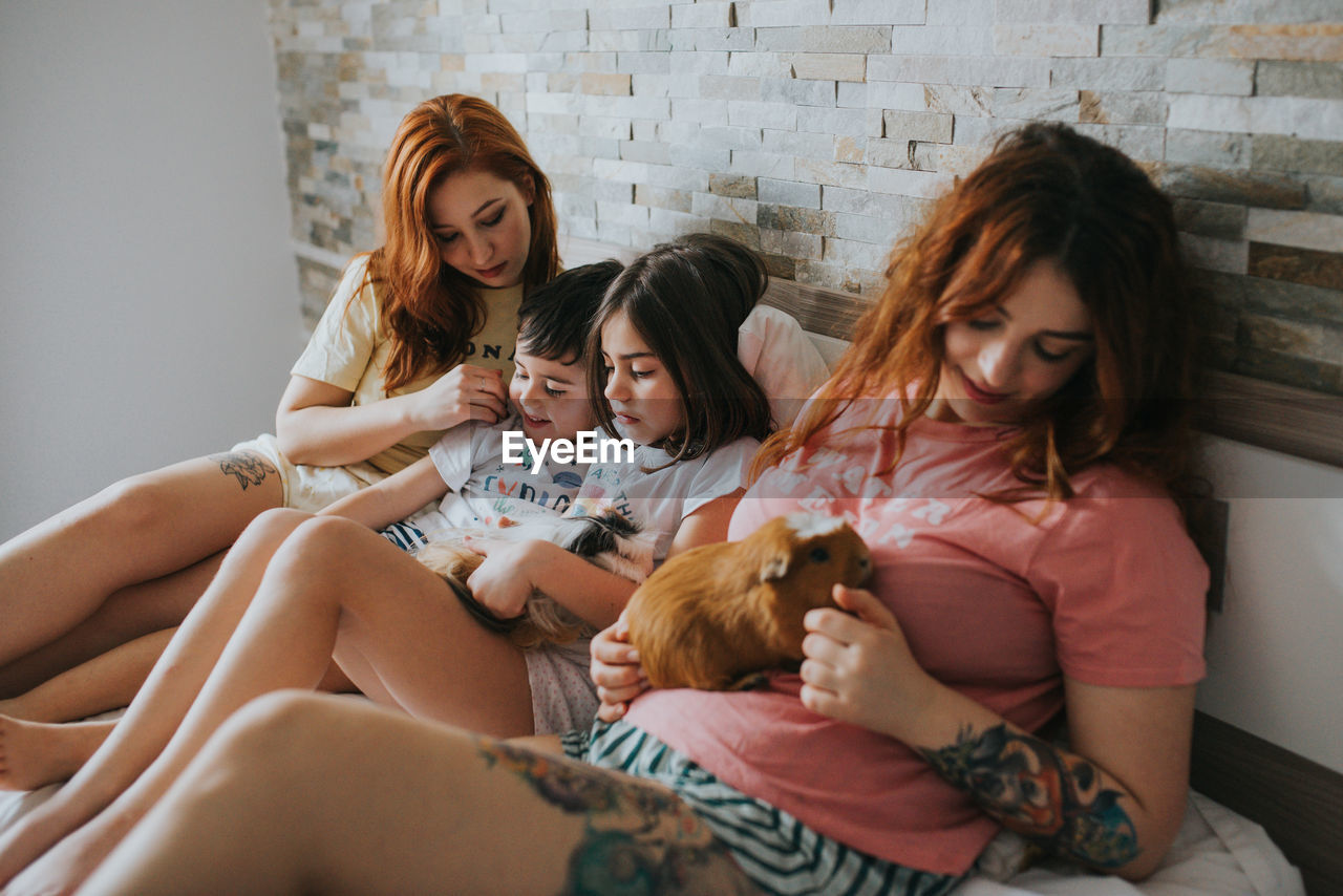 Young same gender parents and kids in pajamas playing with guinea pigs and relaxing on bed in bedroom