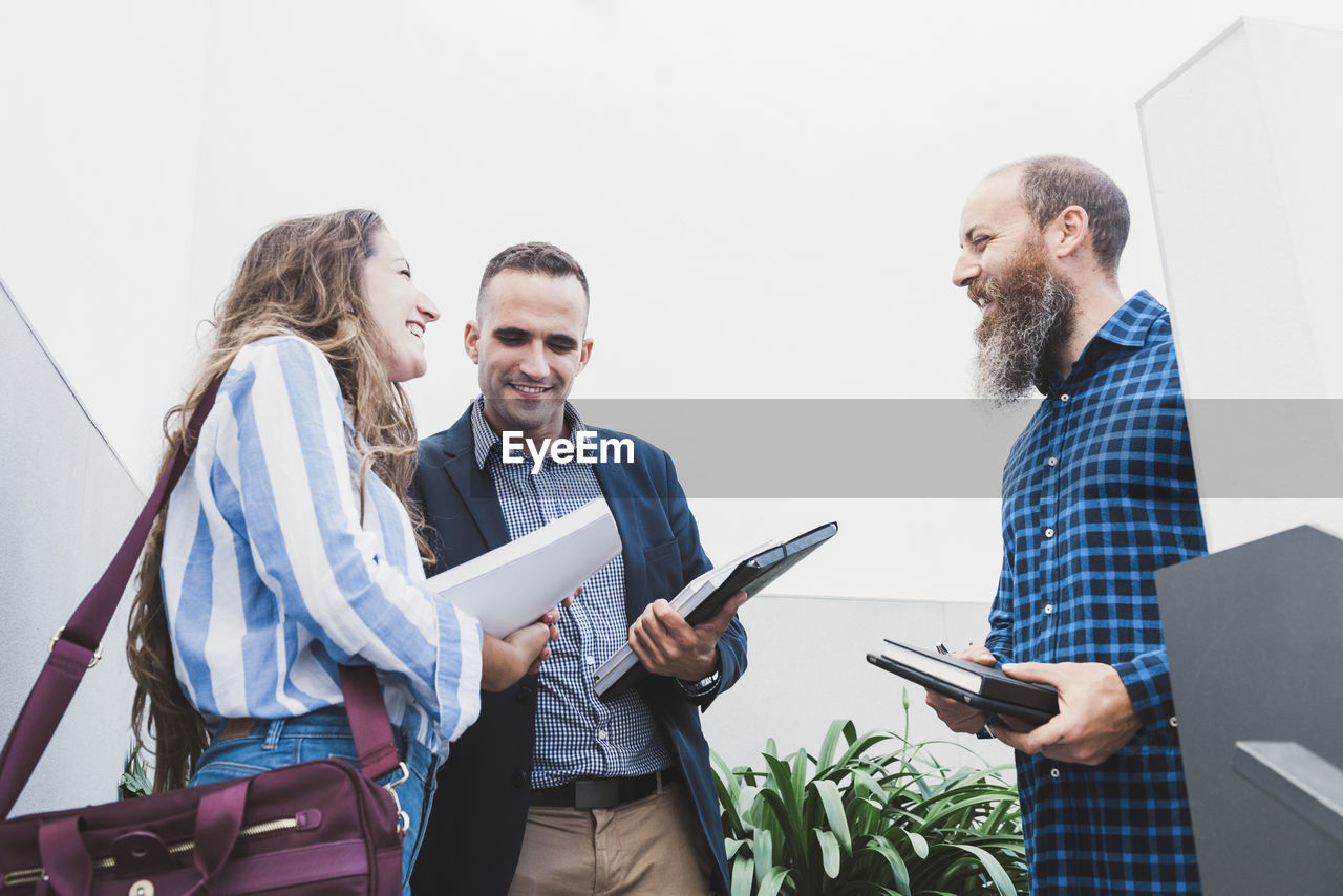 Low angle of group of delighted coworkers standing in modern workplace with plants and discussing project while looking at each other