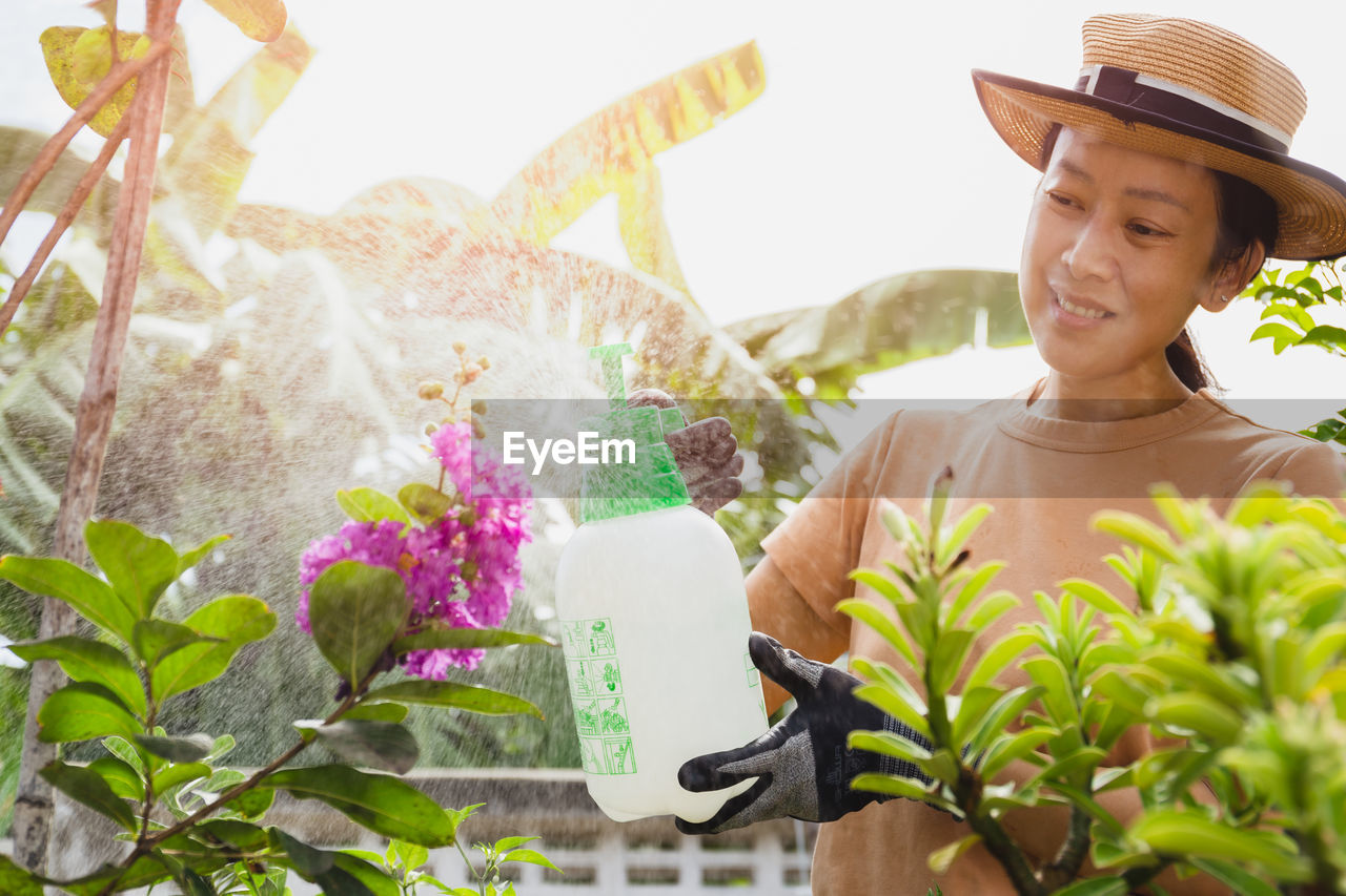 Beautiful asian woman in hat watering plant by foggy spray bottle in garden. 