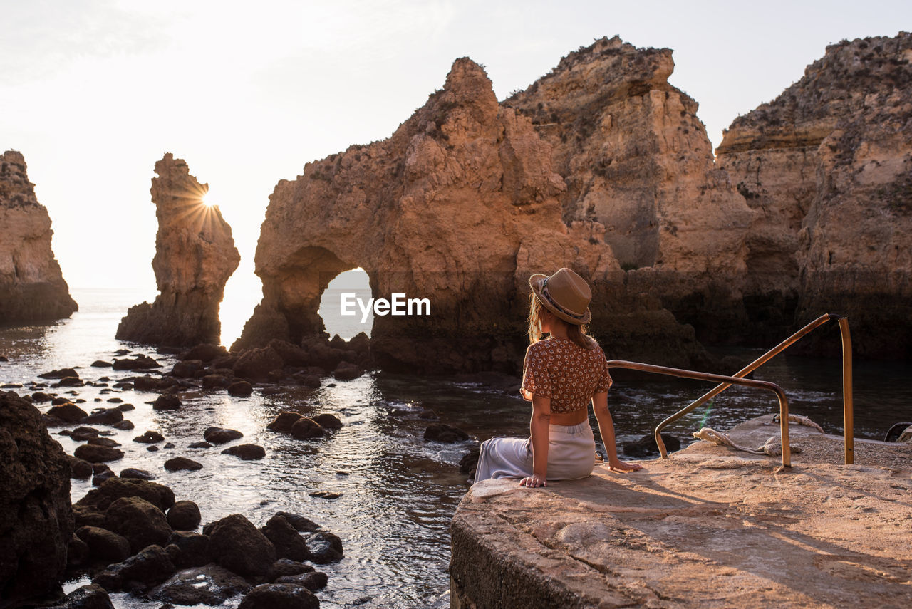 Anonymous female in hat sitting on stone and admiring sea and cliffs at sundown in ponta da piedade in algarve, portugal