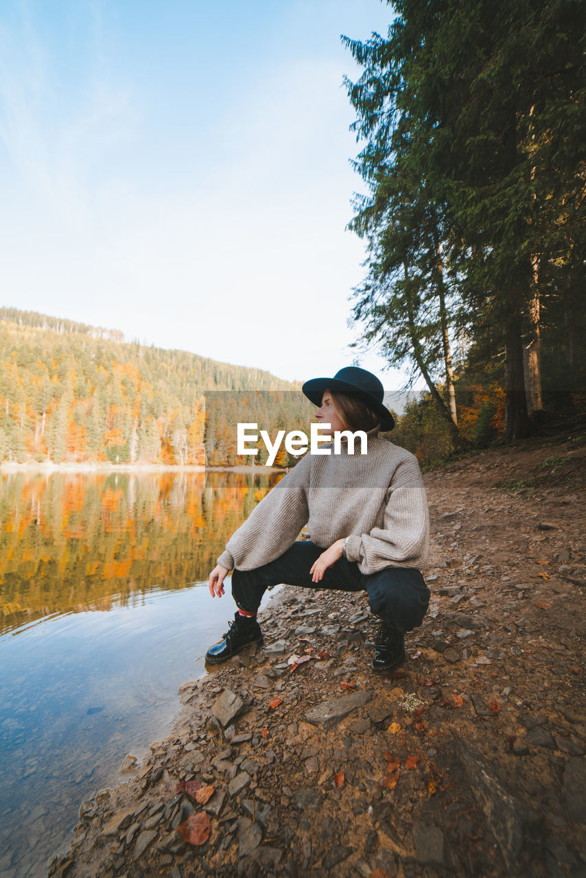 Young cool female tourist in trendy apparel looking away from dry coast against transparent water reflecting autumn trees