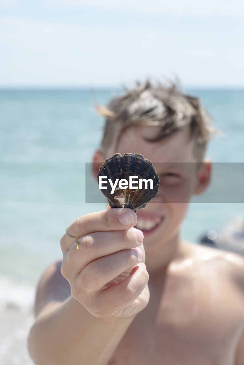 A boy against the background of the sea holds a shell with a hole in his hand. smile. 