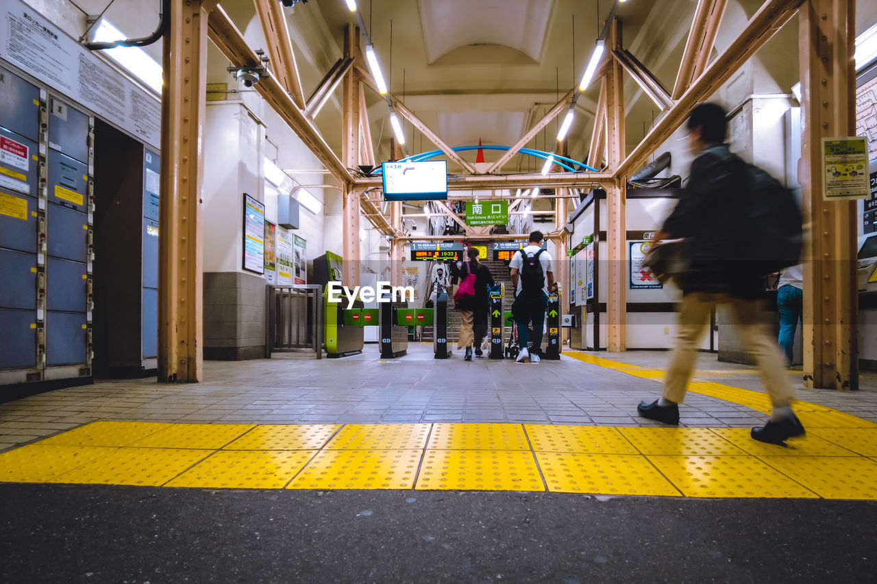 architecture, adult, urban area, full length, transport, public transport, men, infrastructure, walking, city, yellow, transportation, motion, indoors, city life, built structure, business, rear view, person, travel, lifestyles, street, one person, store, women, road, blurred motion