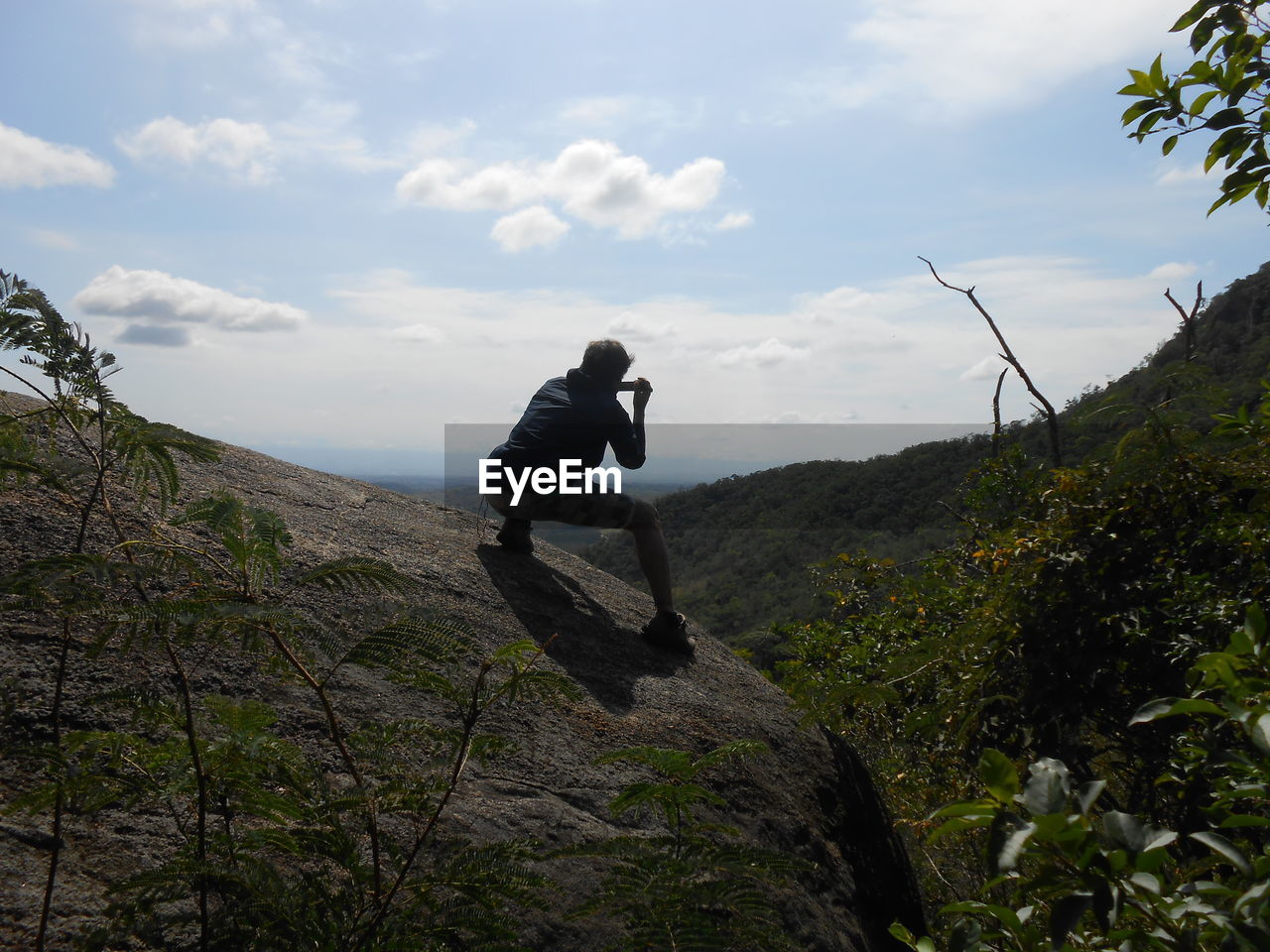 Rear view of man crouching on cliff against sky