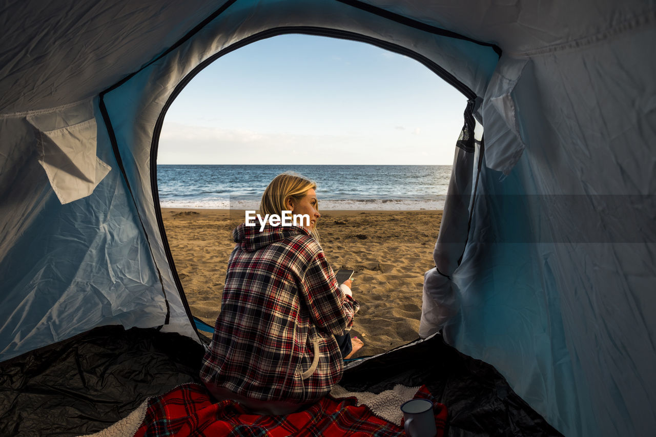 Woman sitting in tent at beach against sky