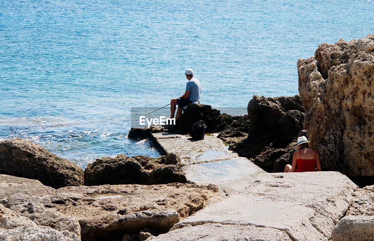 REAR VIEW OF MAN STANDING ON ROCK BY SEA