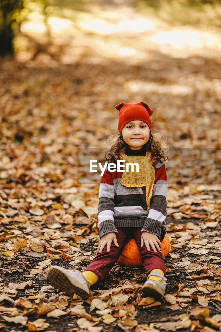 Happy cute little girl child in warm bright clothes sitting on a pumpkin in the autumn park outdoors