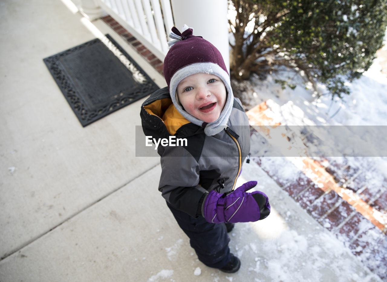Boy in winter gear standing on snowy front porch looks up at camera