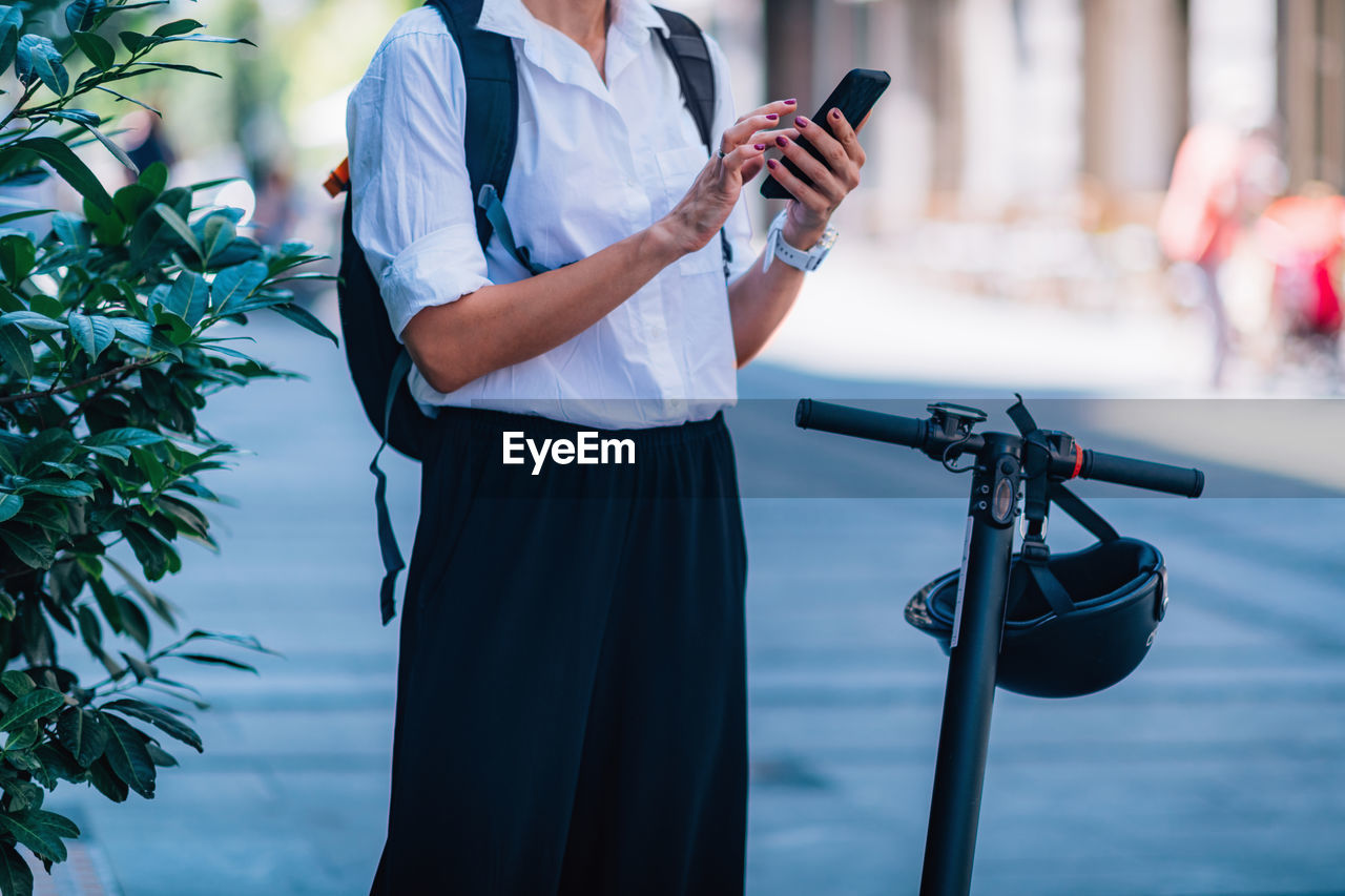 Midsection of woman using phone while standing with scooter on road