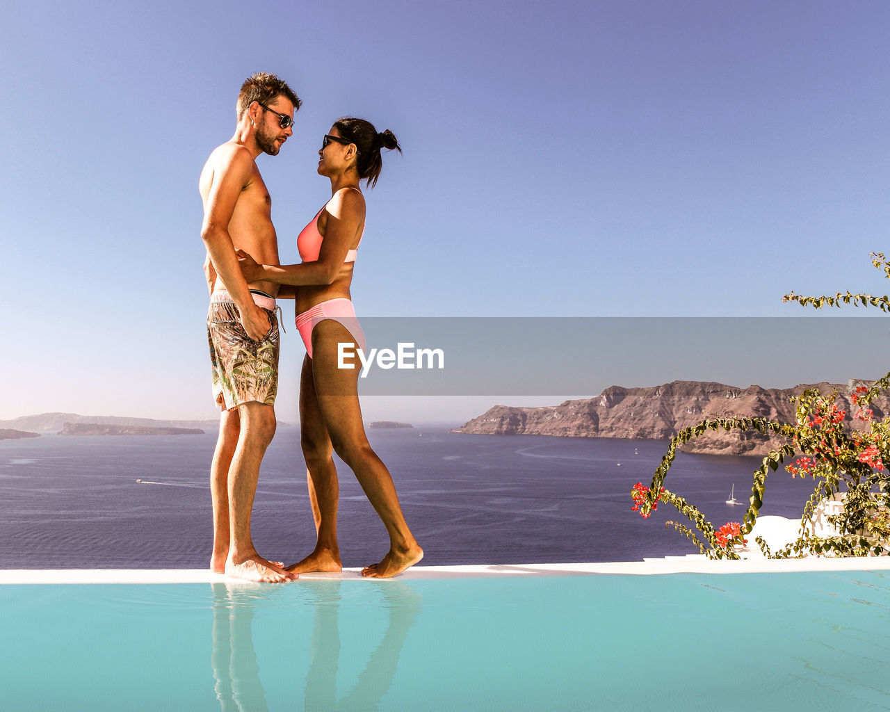 Full length of young couple standing by infinity pool at sea against clear sky