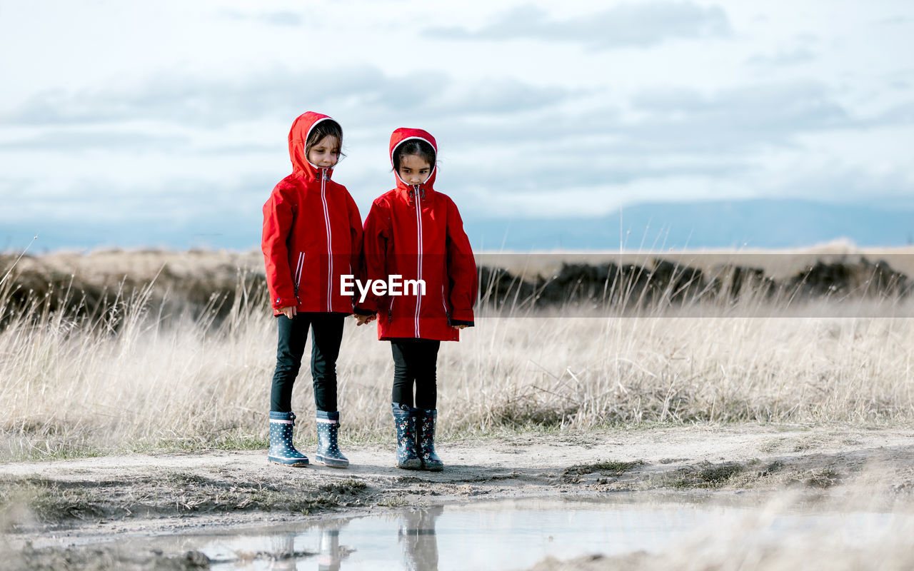 Carefree kids in raincoats and rubber boots in puddle on sunny day in countryside