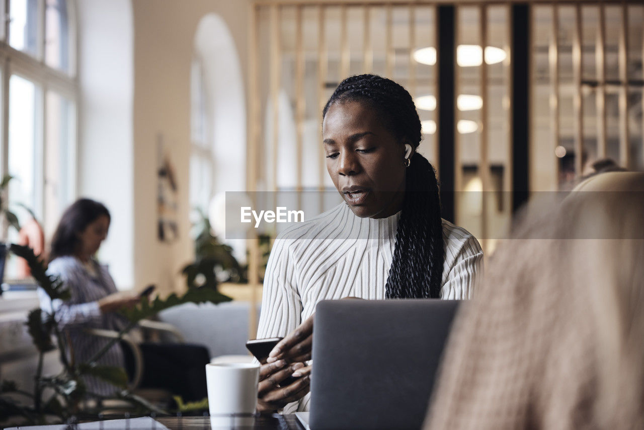 Businesswoman talking through wireless in-ear headphones and using smart phone while sitting at coworking office