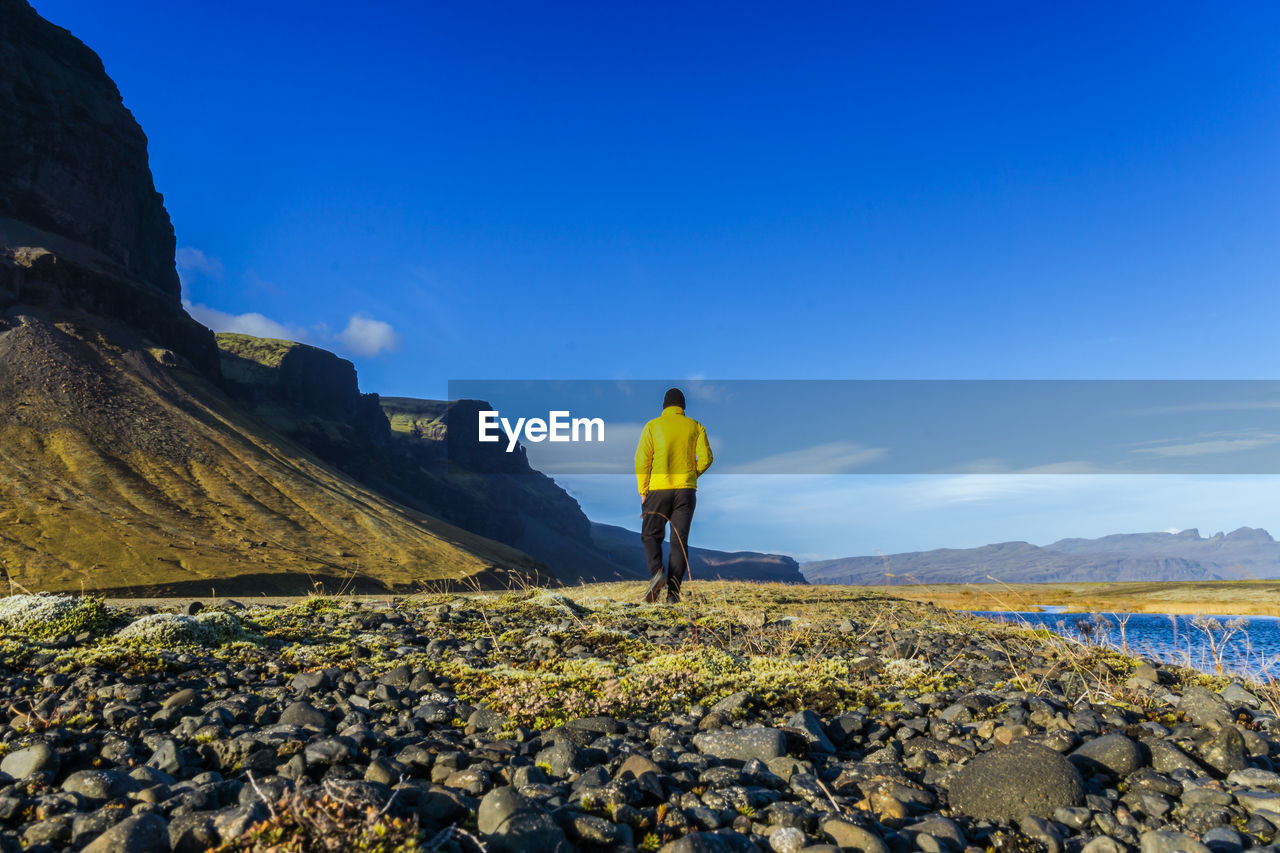 Rear view of hiker walking on lakeshore by mountains against sky