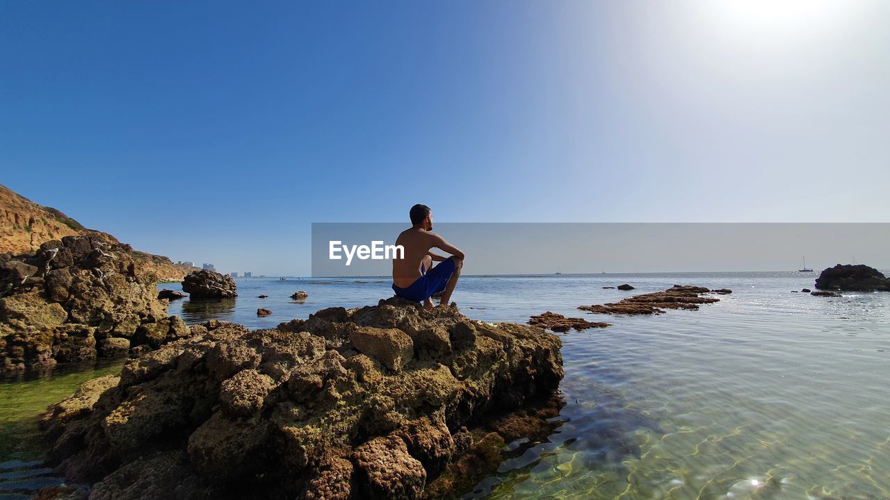 Man standing on rock by sea against clear blue sky