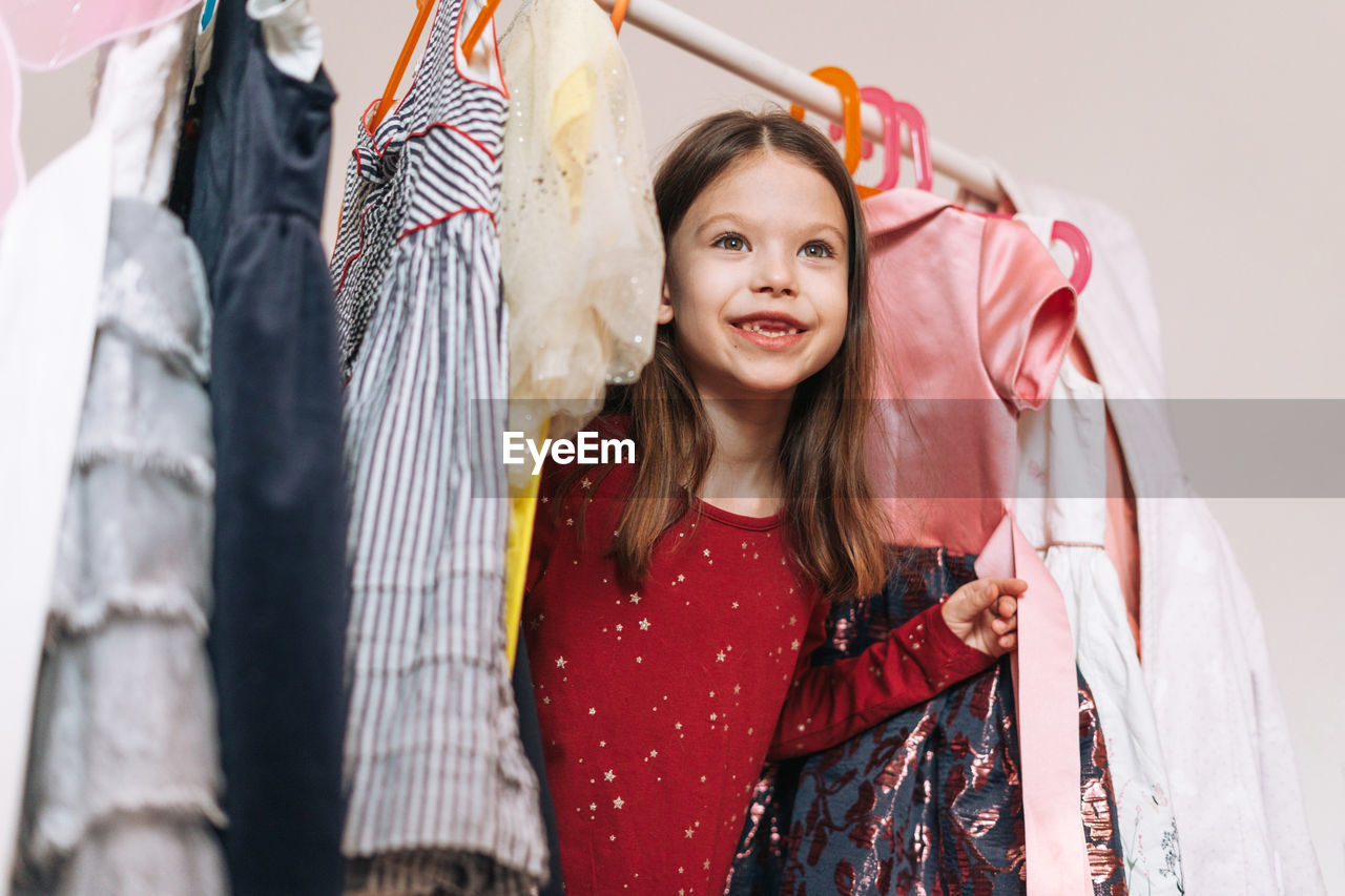 Smiling girl with long dark hair in red dress among dresses in wardrobe in children room at home