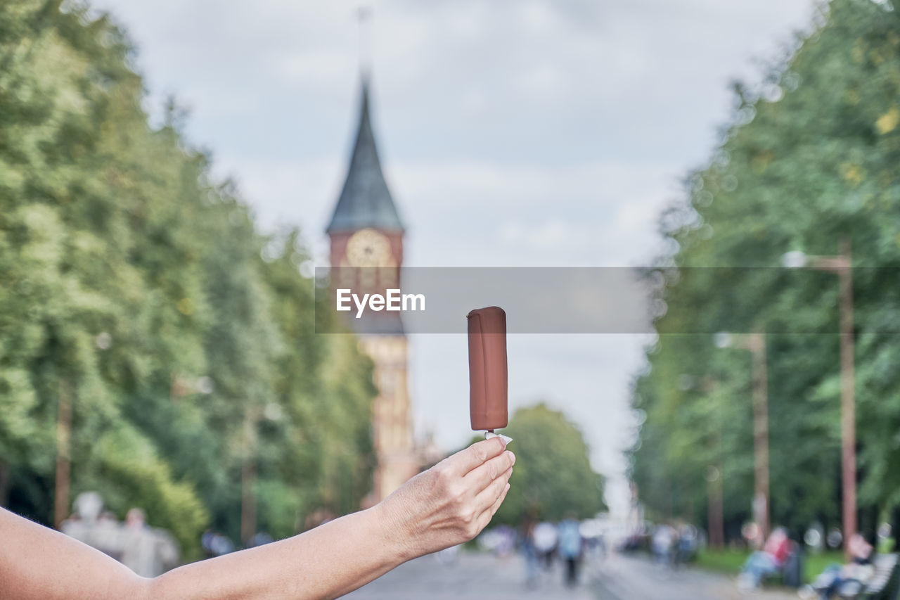 Senior woman's hand holding ice cream in chocolate over background of park and cathedral 