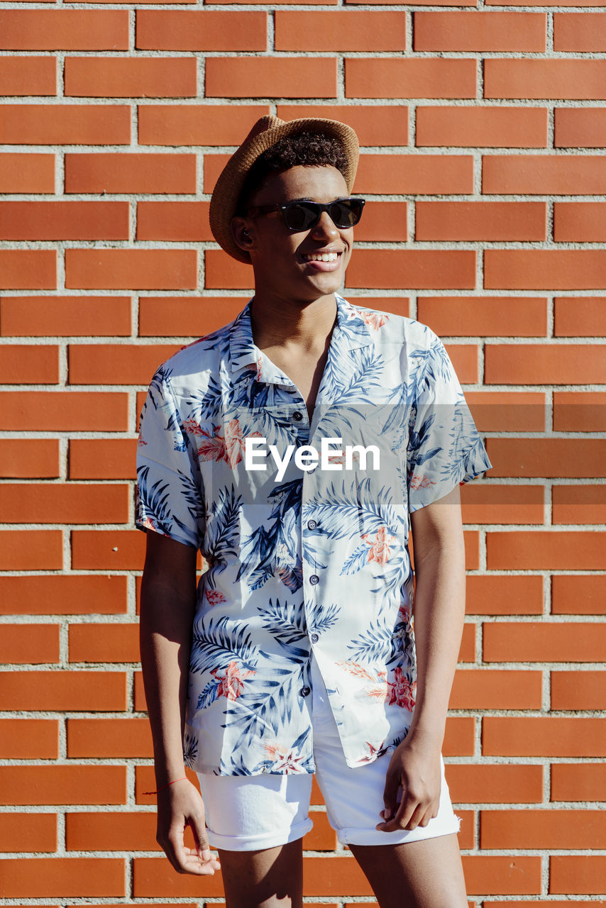 Trendy young black man with sunglasses and a hat posing on the street next to a brick wall