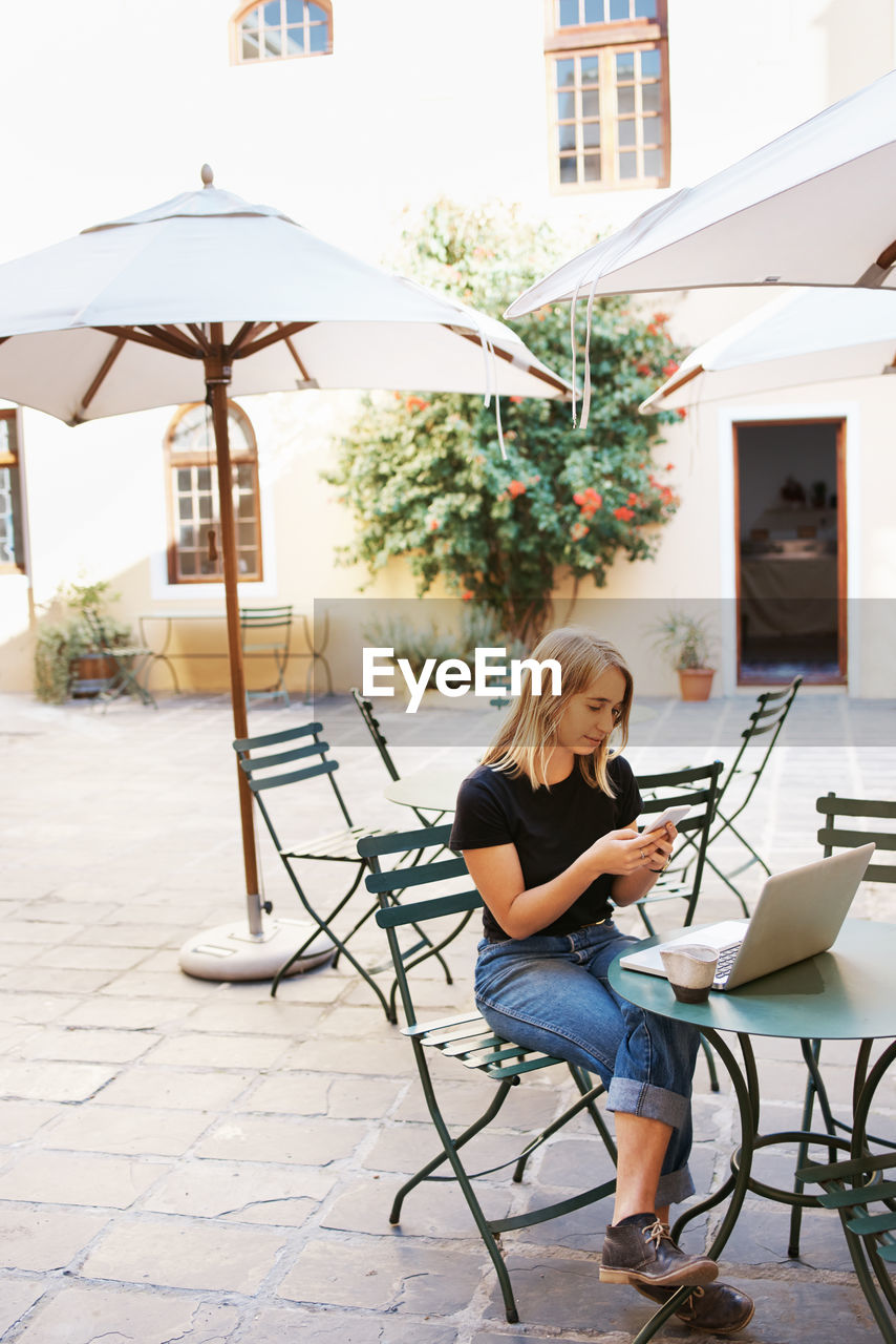 Young woman using mobile phone while sitting on chair at sidewalk cafe