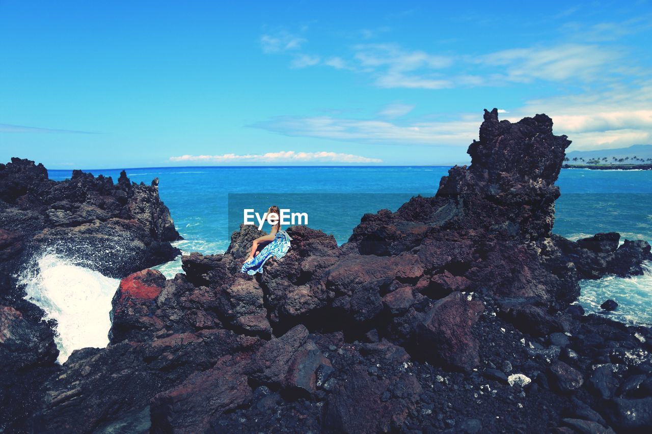 Side view of young woman sitting rock formations by sea against blue sky