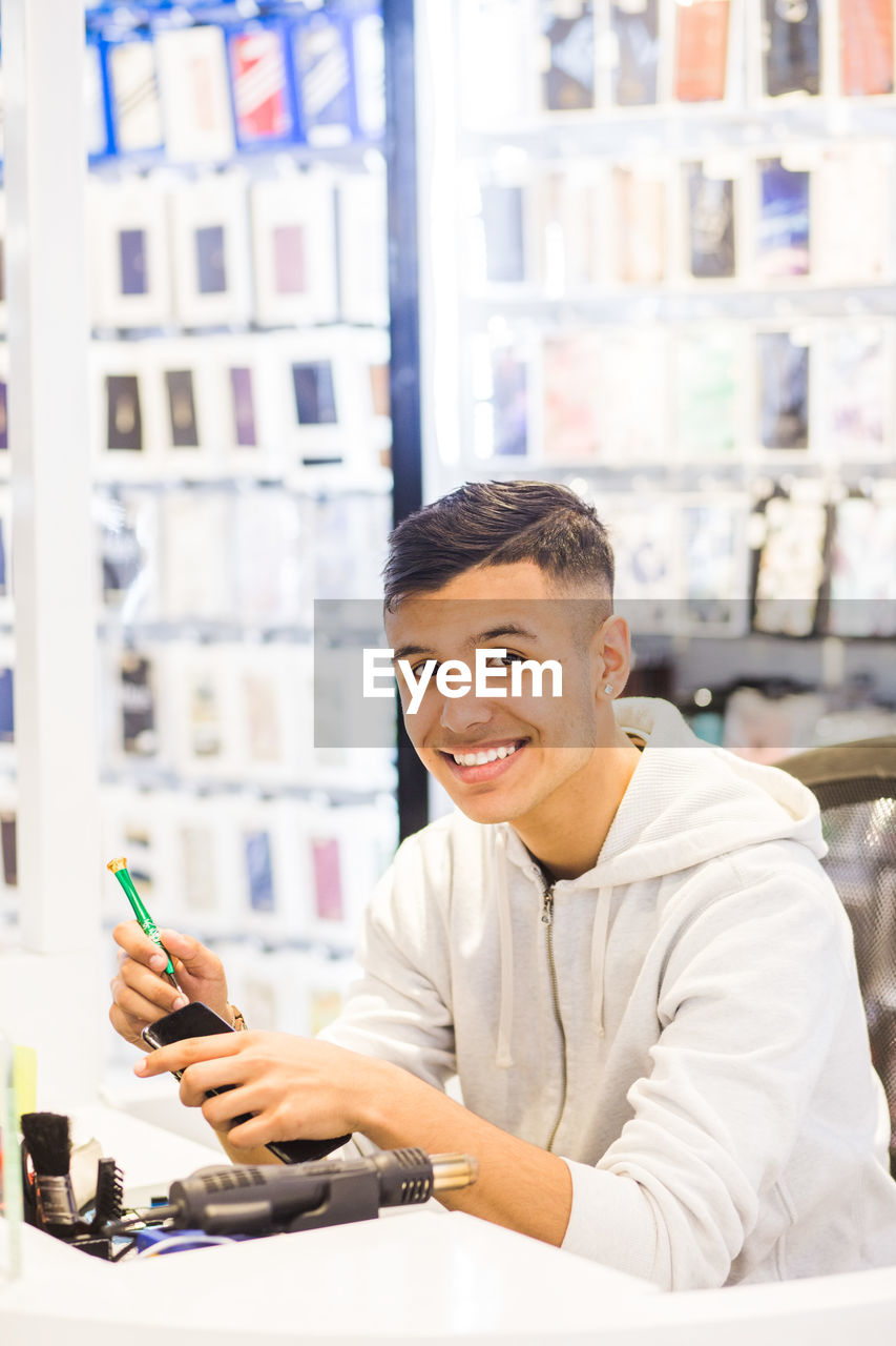 Portrait of smiling teenage trainee repairing mobile phone while sitting at illuminated desk in store