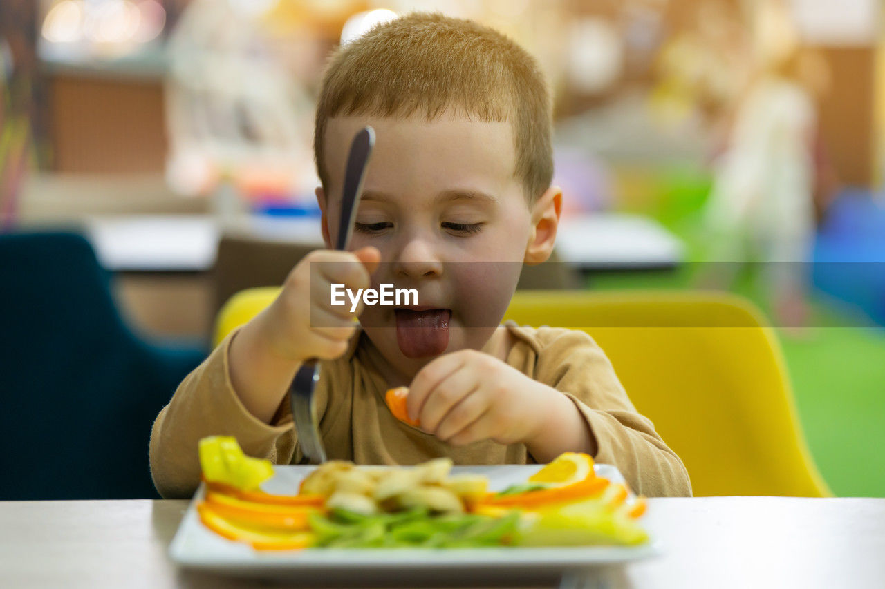 A funny cute three year old toddler boy eats fresh fruit while sitting at a table in a public place.