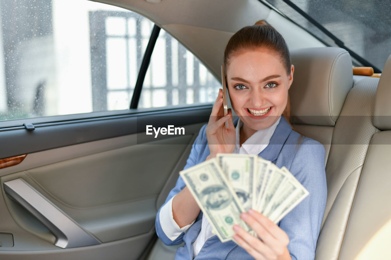 Portrait of smiling businesswoman with paper currency talking on phone while sitting in car