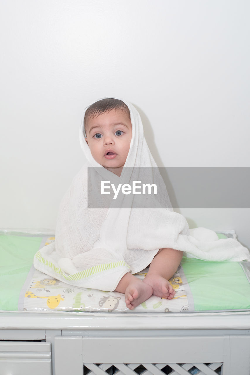 Portrait of cute baby boy wrapped in towel while sitting on bed against wall