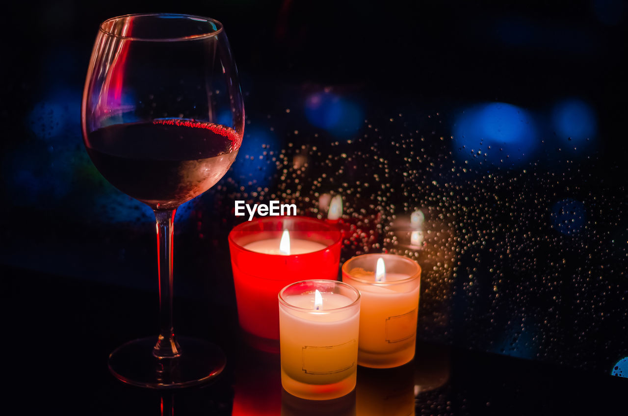 A glass of red wine with burning candles put beside window that have rain drop on dark background.