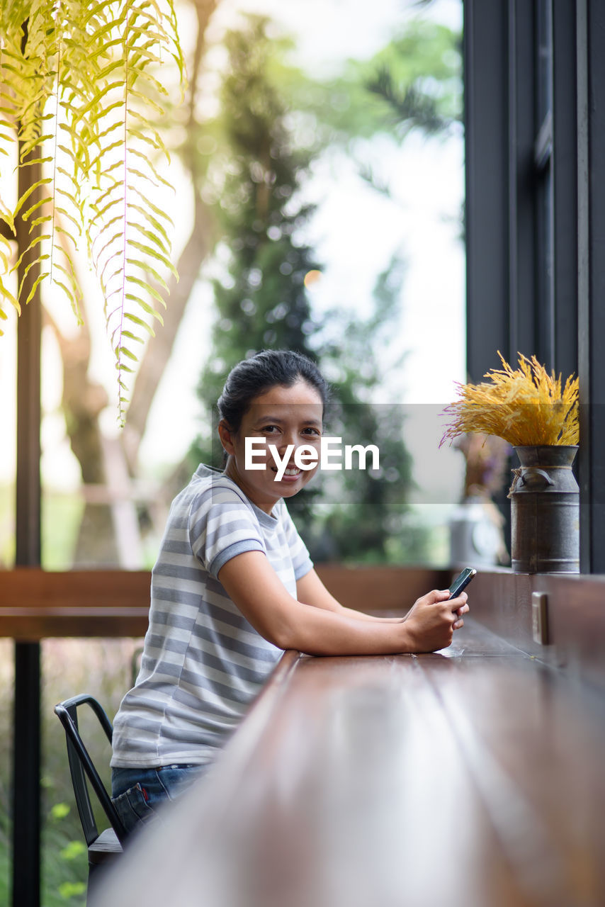 Portrait of smiling woman sitting with mobile phone at table by window