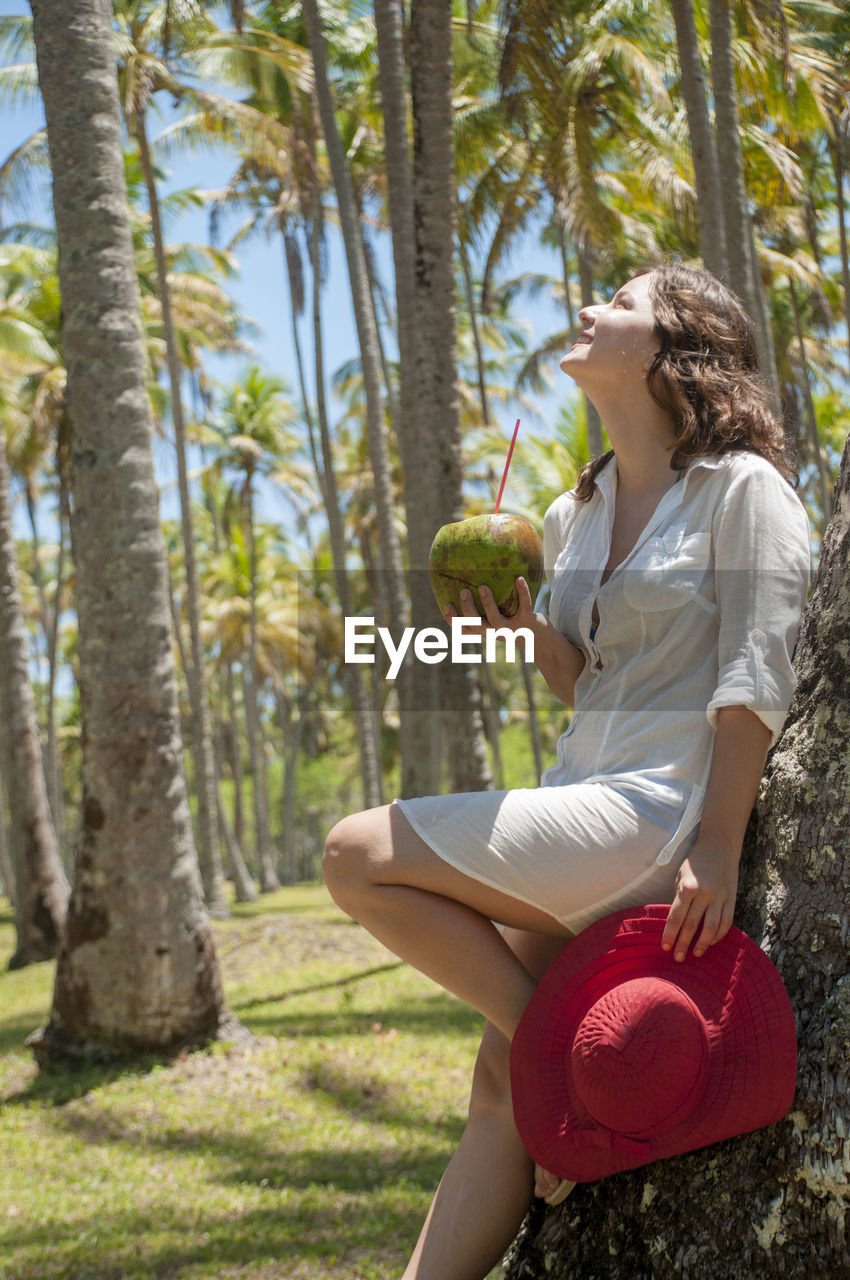 Smiling woman with coconut leaning on palm tree