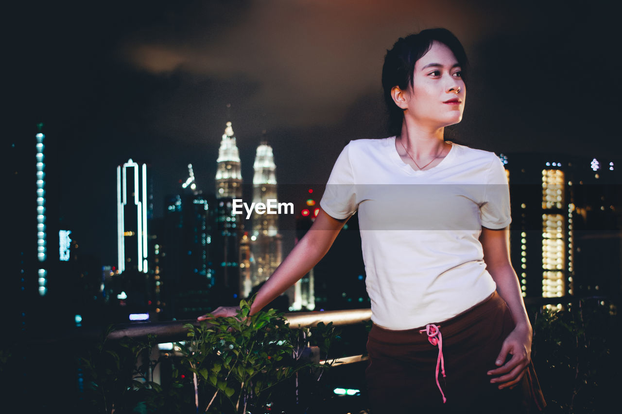 Young woman looking away while standing against illuminated buildings in city at night