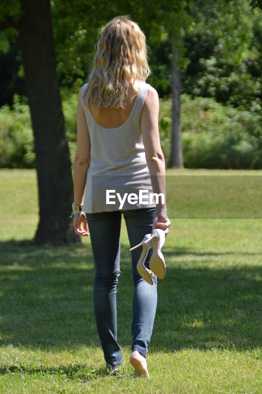 Rear view of woman holding high heels walking on grass at park