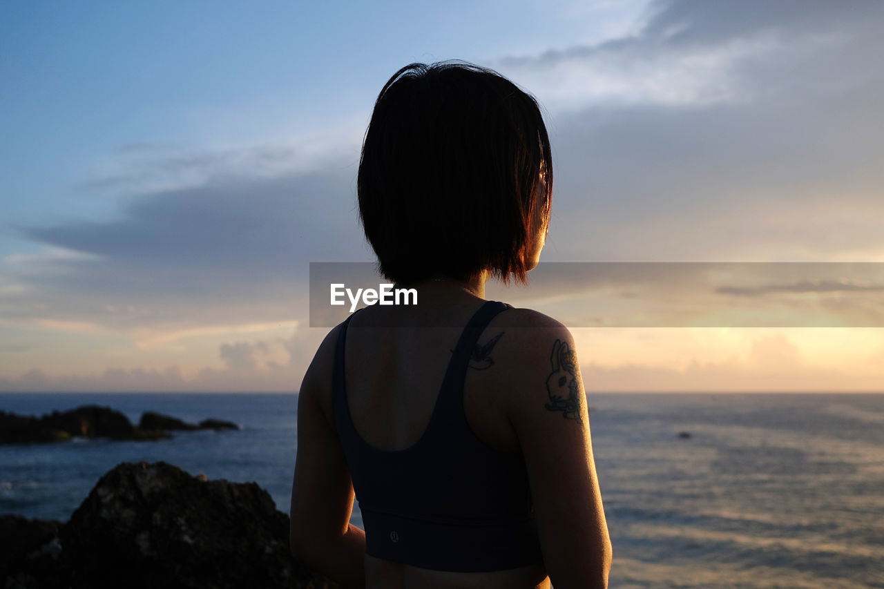 rear view of young woman standing at beach against sky during sunset