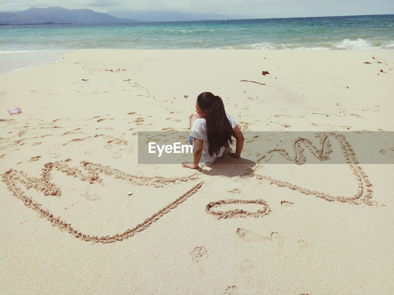 Rear view of woman sitting by wings drawing on sand at beach