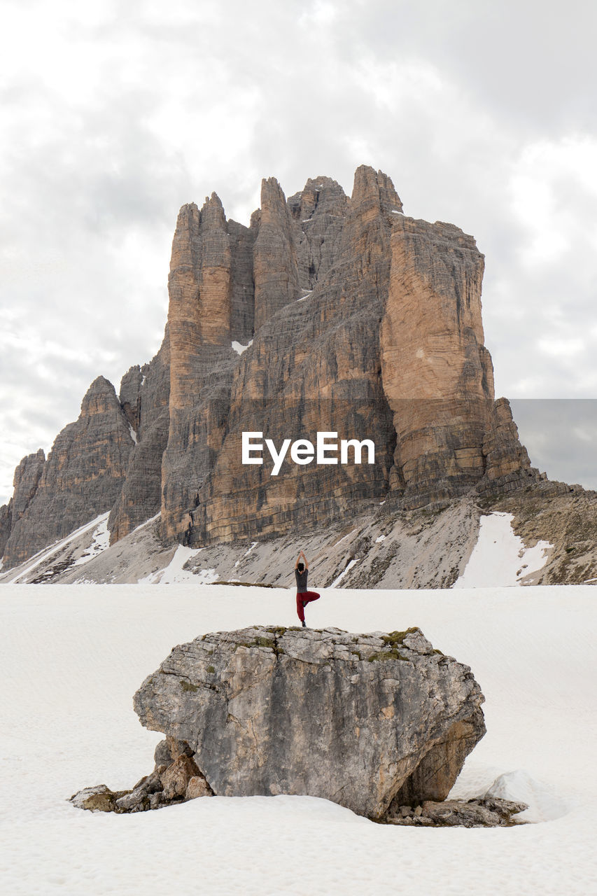 Remote view of traveler standing in tree pose on rock and practicing yoga in snowy dolomites mountains