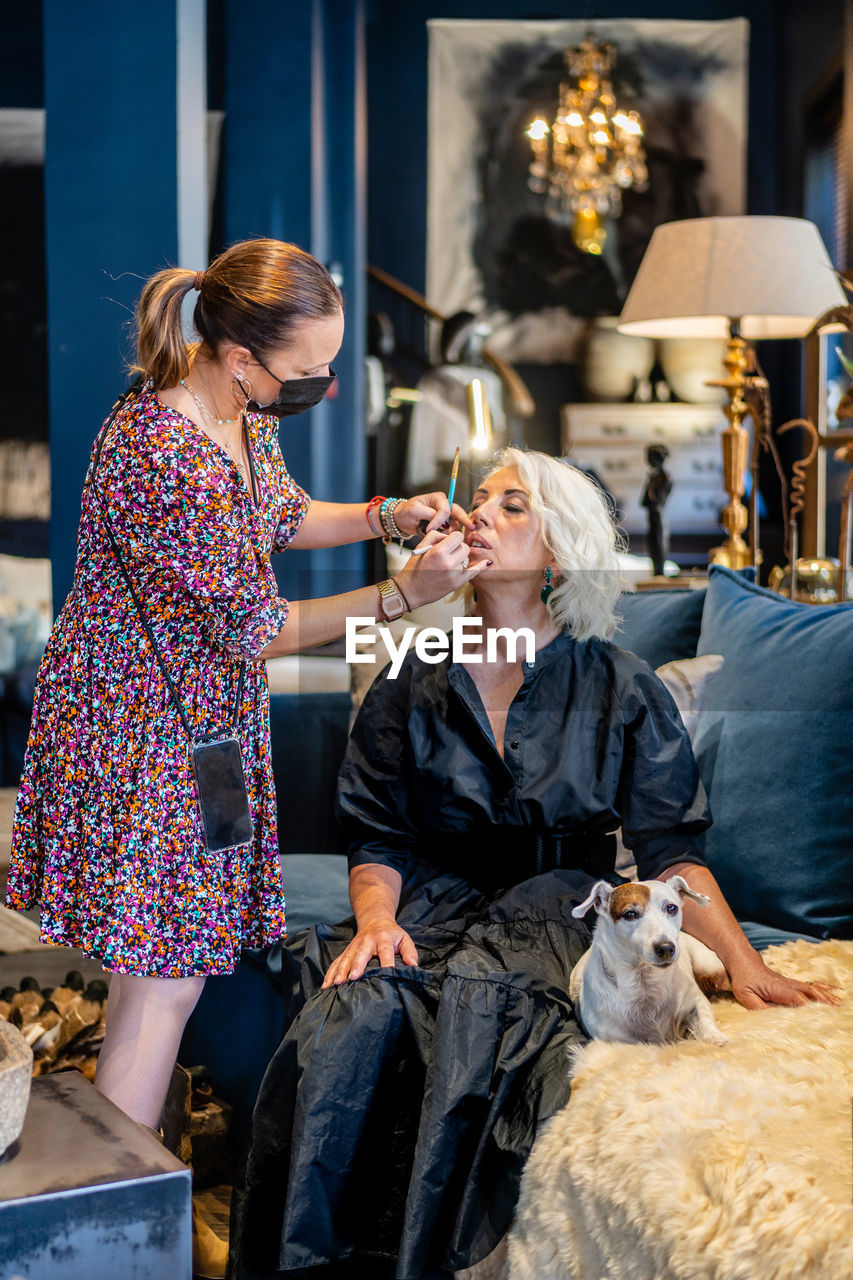 Cosmetician in protective mask applying makeup to elegant mature lady sitting on sofa with purebred dog in stylish living room at home