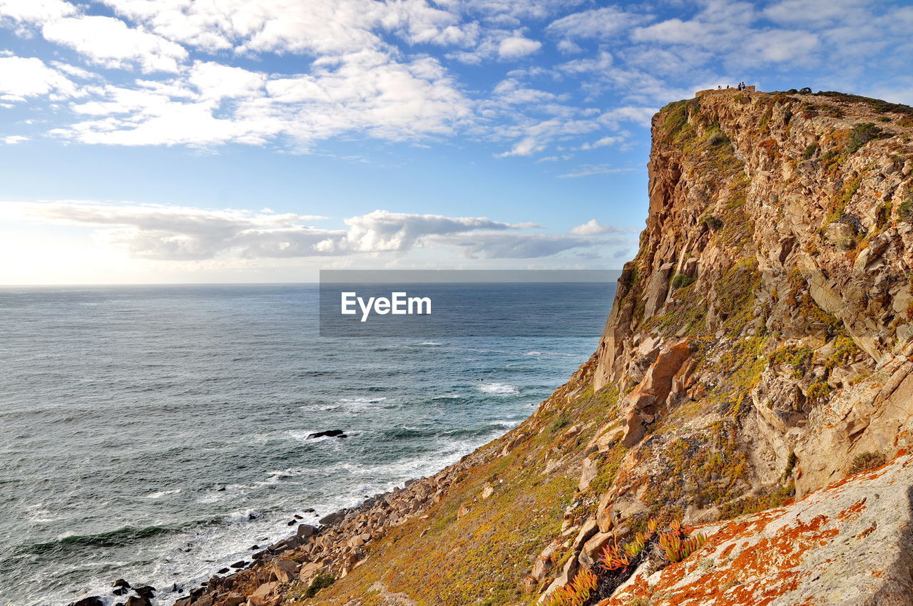 Scenic view of sea by rock formation against cloudy sky during sunny day